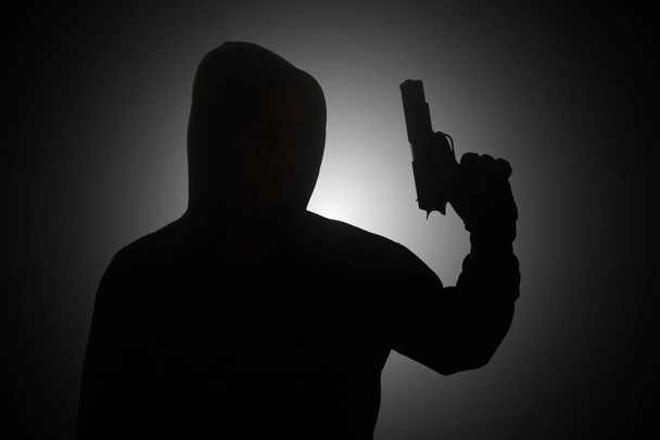 Hombre misterioso con capucha negra sosteniendo una pistola, disparando con un arma. Silueta e imagen concepto oscuro - Foto, imagen