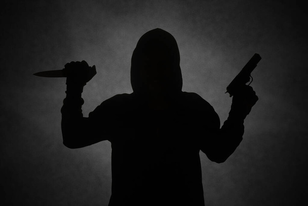 Μυστηριώδης άντρας φορώντας μαύρη κουκούλα κρατώντας ένα πιστόλι, πυροβολώντας με ένα όπλο κρατώντας ένα μαχαίρι. Σιλουέτα και σκοτεινή εικόνα έννοιας - Φωτογραφία, εικόνα