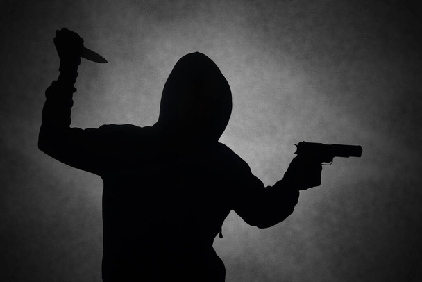 Salaperäinen mies, jolla on musta huppari, pitelee pistoolia, ampuu aseella pitäessään veistä. Siluetti ja tumma konsepti kuva - Valokuva, kuva