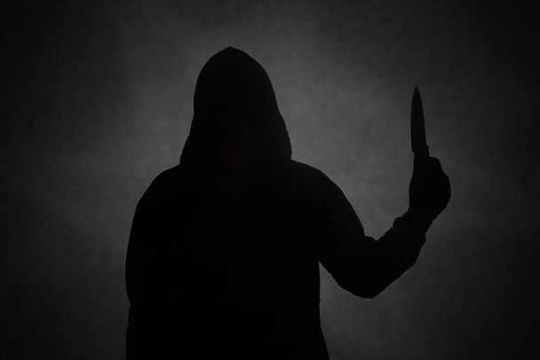 Μυστηριώδης άντρας που φοράει μαύρη κουκούλα κρατώντας ένα μαχαίρι για να μαχαιρώσει κάποιον. Έννοια του εγκλήματος και της εγκληματικότητας - Φωτογραφία, εικόνα