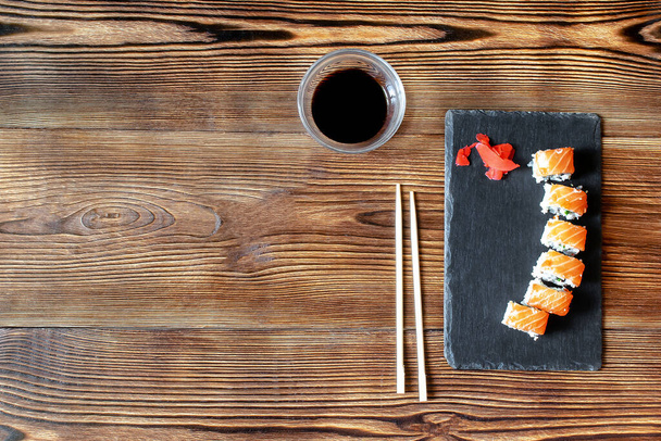 Rollos de sushi de pescado con salmón, jengibre, salsa de soja y palillos sobre tabla de servir de corte negro sobre fondo rústico de madera con espacio para copiar. Mariscos, Servicio de comidas desde el espacio de copia de concepto de restaurante. - Foto, imagen