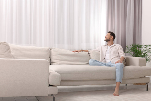 Happy man on resting sofa near window with beautiful curtains in living room. Space for text - Foto, Imagem