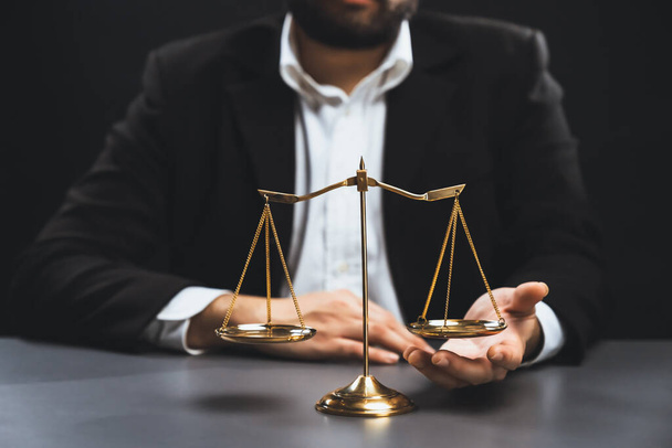 Asianajaja mustassa puvussa istuu tarkkaavaisesti toimistopöydällään, jossa on kultainen vaaka tasapaino, oikeudellisen oikeudenmukaisuuden ja koskemattomuuden symboli, tasapainoinen ja eettinen päätöksenteko tuomioistuimessa. tasavertaisuus - Valokuva, kuva