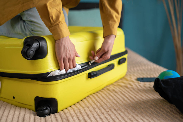An unrecognizable young woman getting ready for the trip, vacation, travel, presses her foot on a crowded suitcase to close it. Packing and trying to close suitcase on bed. Close-up - Foto, Imagen