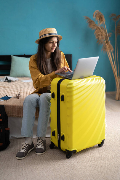 Traveler γυναίκα κάθεται χαμογελώντας στο κρεβάτι στην κρεβατοκάμαρα με αποσκευές τρόλεϊ, ενώ χρησιμοποιώντας το φορητό υπολογιστή κρατήσεις διακοπές, καλοκαιρινό ταξίδι σε απευθείας σύνδεση. Αντιγραφή χώρου - Φωτογραφία, εικόνα