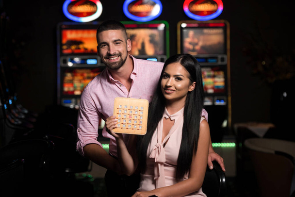 Portrait of a Young Beautiful and Well Dressed Couple Playing in the Casino and Celebrate Holding Bingo Wood Game Card - Fotografie, Obrázek