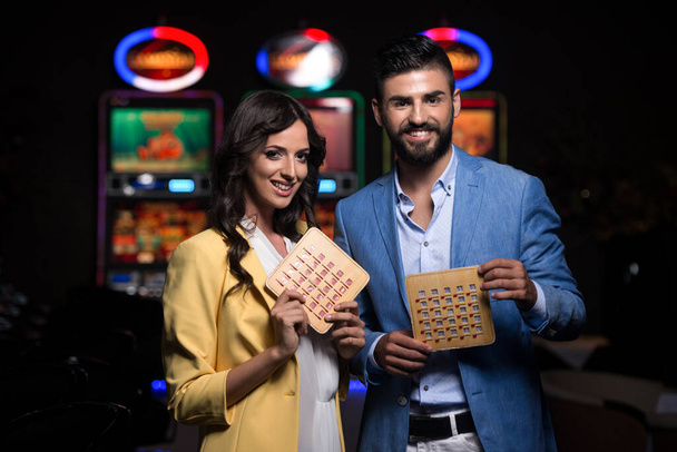 Portrait of a Young Beautiful and Well Dressed Couple Playing in the Casino and Celebrate Holding Bingo Wood Game Card - Zdjęcie, obraz