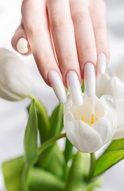 Female hand with spring nail design. White nail polish manicure. Female model hand with perfect manicure hold white tulip flowers on white silk background - Photo, Image