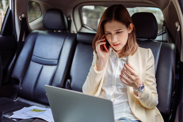 Bizneswoman rozmawia przez telefon komórkowy i używa laptopa w samochodzie. - Zdjęcie, obraz