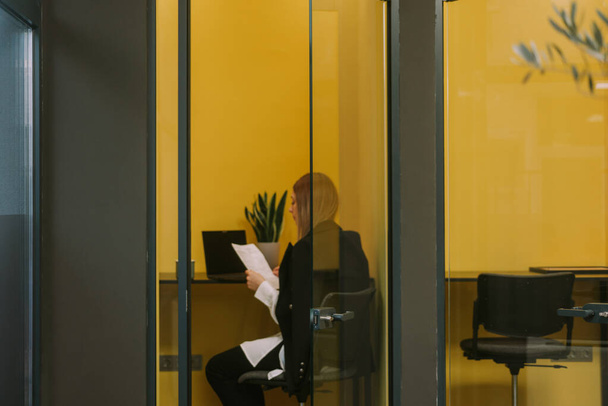 Sarışın bir iş kadını çalışma alanındaki küçük sarı odada otururken dosyalara bakıyor. - Fotoğraf, Görsel