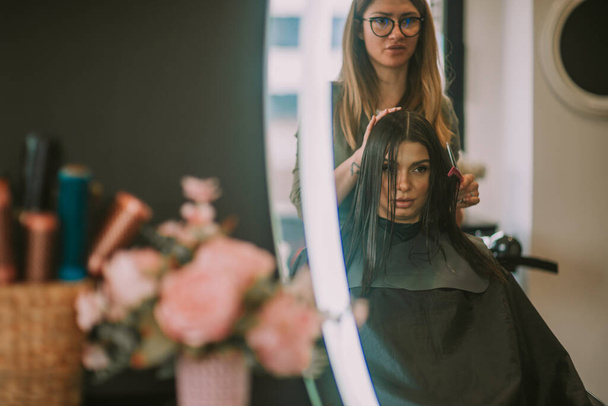 Blondynka fryzjer szczotkowanie swoich klientów proste brązowe włosy podczas gdy ona siedzi w salonie fryzjerskim - Zdjęcie, obraz