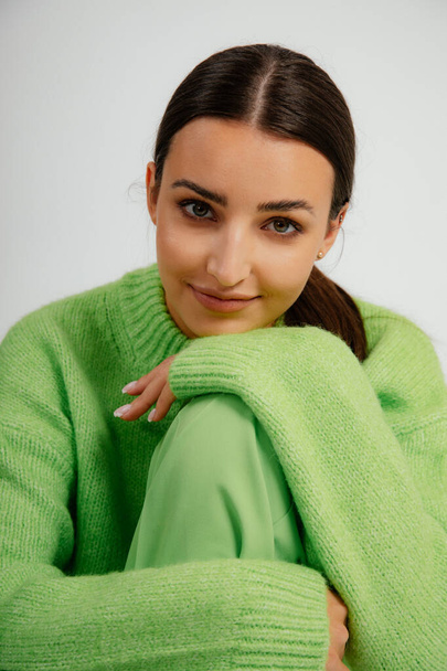 Verticaal positieve, vrolijke, moderne brunette in groene trui, kijkend naar camera met glimlach en leunend op knie door handen met manicure op witte studio achtergrond. Natuurlijke make-up en schoonheid - Foto, afbeelding