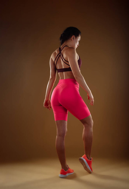 Donna sexy in pantaloncini rosa e top sportivo bordeaux in posa in studio. Indietro vista foto - Foto, immagini