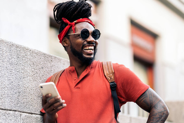 Stockfoto van een aantrekkelijke Afro-Amerikaanse jongen met dreadlocks en een bandana met zijn telefoon. Hij lacht.. - Foto, afbeelding
