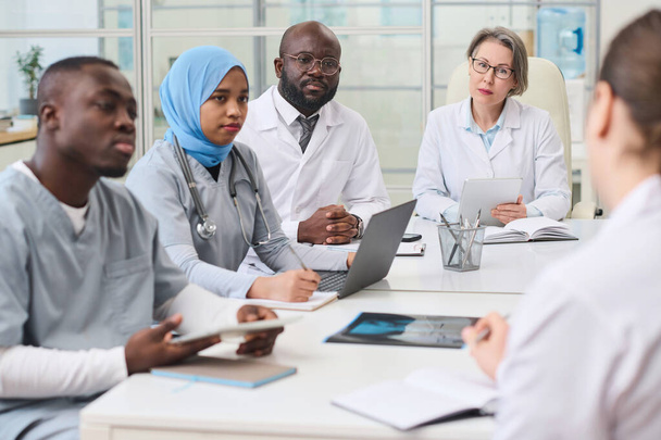 Group of doctors listening to their colleague while sitting at table during meeting in office, they discussing difficult operation or disease - Foto, Imagen