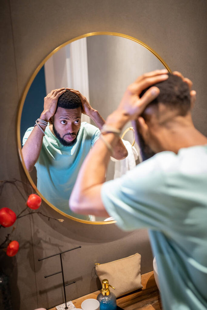 Miesten hiustenlähtö. Järkyttynyt nuori afroamerikkalainen mies katselee kylpyhuoneen peiliin koskettaen hiuksia, masentunut musta mies murehtimassa kaljuuntumista. Mies kaljuuntuminen ja mielenterveyden käsite - Valokuva, kuva