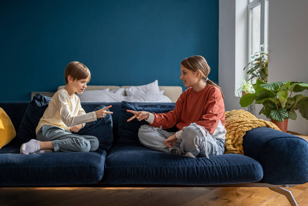 Healthy sibling relationships. Playful children brother and sister sitting on sofa playing rock-paper-scissors game during leisure time, bonding together at home. Sibling play concept - Φωτογραφία, εικόνα