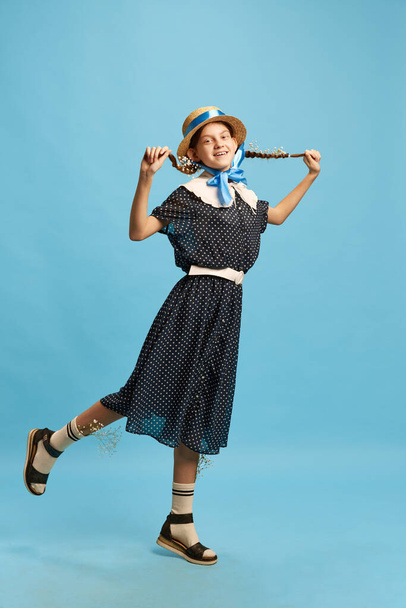 Flores en coletas. Una joven linda chica con vestido y sombrero posando con emociones positivas sobre fondo de estudio azul. Concepto de moda moderna, singularidad, estado de ánimo, fashionista, arte, anuncio - Foto, Imagen