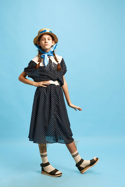 Arrogant little girl. One pretty young girl wearing vintage dress and hat looking at camera with dislike face over blue background. Vintage, retro, uniqueness, art, childhood, ad concept - Photo, image