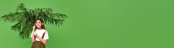 Durante la lluvia. Una linda niña de pie con la hoja de la planta en su lugar paraguas sobre el fondo verde. Banner con espacio de copia. Concepto de belleza, emociones humanas, estado de ánimo, arte, moda moderna, anuncio - Foto, Imagen