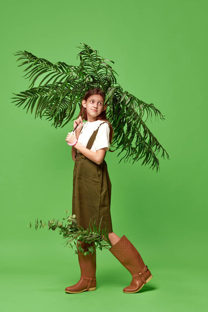 Rainy season. Charming young girl wearing boots standing with palm instead of umbrella over green background. Concept of beauty, human emotions, mood, art, modern fashion, ad - Photo, Image