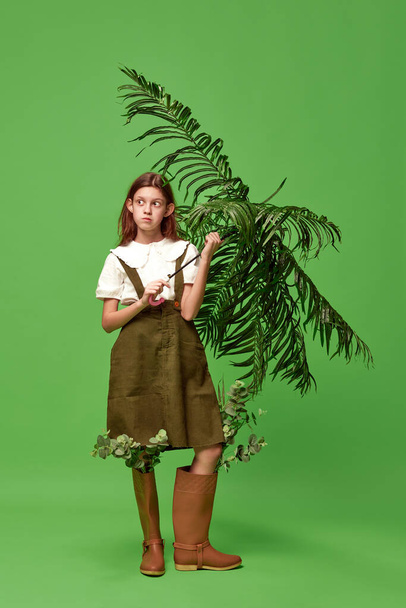 Garden worker. Little pretty girl wearing boots standing with palm leaf instead of umbrella over green background. Concept of nature, beauty, human emotions, mood, art, modern fashion, ad - Photo, Image
