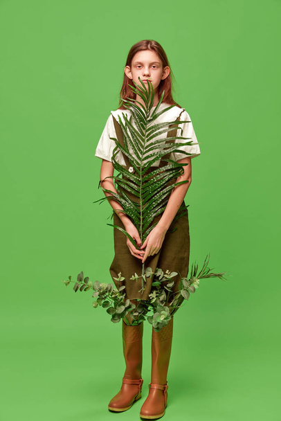 Child of nature, forest. One pretty girl with long hair wearing boots holding palm leaf over green background. Concept of beauty, human emotions, creative, mood, art, modern fashion, ad - Photo, Image