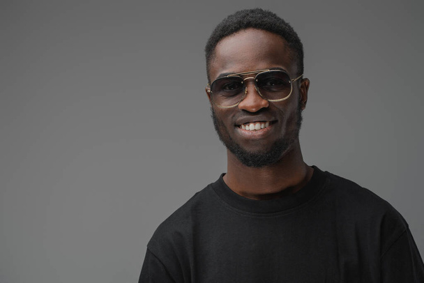 Studio zdjęcie uśmiechniętego faceta afrykańskiego etnicznego ubranego w modne okulary przeciwsłoneczne i strój casual. - Zdjęcie, obraz