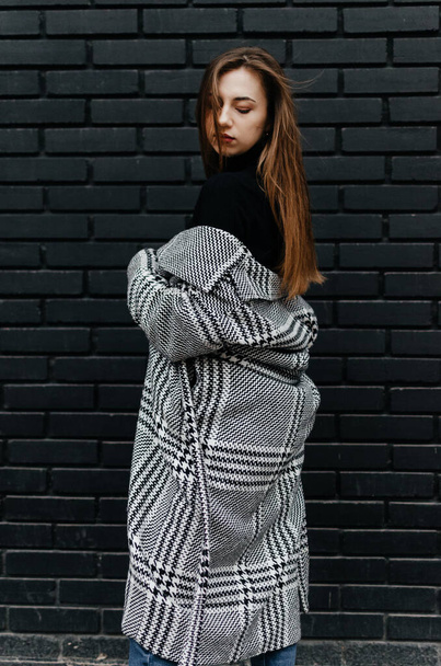 A woman in a coat stands in front of a black brick wall - Foto, Imagen