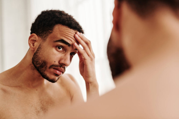 Jonge knappe man onderzoekt rimpels op zijn gezicht terwijl hij naar de spiegel kijkt - Foto, afbeelding