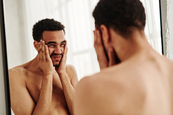 Young man holding his head in hands and looking at mirror with displeasure - Photo, Image