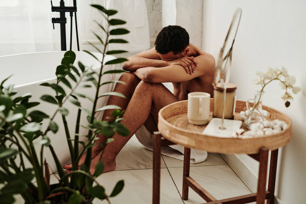 Молодий чоловік сидить на підлозі у ванній і плаче, ховаючи обличчя в руках
 - Фото, зображення