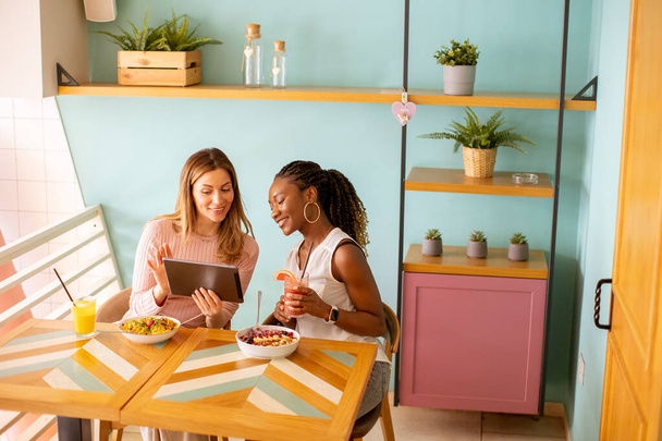 Kaksi kaunis nuori musta ja valkoihoinen nainen, jolla on hauskaa, juo tuoreita mehuja ja ottaa terveellisen aamiaisen kahvilassa - Valokuva, kuva