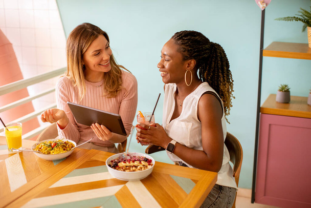 Kaksi kaunis nuori musta ja valkoihoinen nainen, jolla on hauskaa, juo tuoreita mehuja ja ottaa terveellisen aamiaisen kahvilassa - Valokuva, kuva