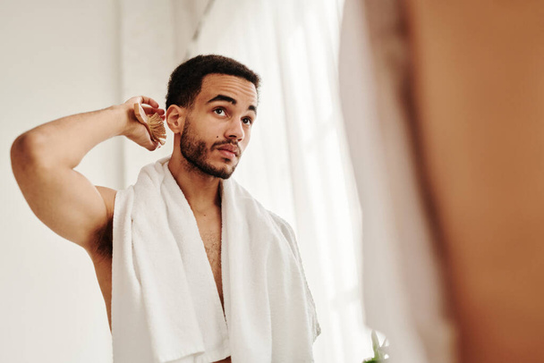 młody przystojny mężczyzna szczotkowanie jego włosy stojąc przed lustrem po prysznic w łazience - Zdjęcie, obraz