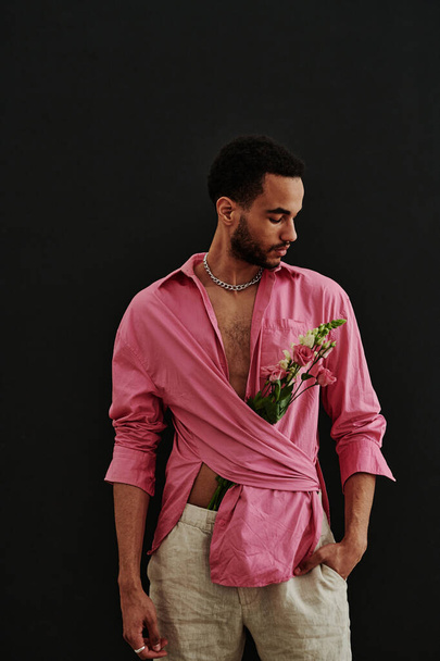 Retrato de hombre de raza mixta guapo en camisa rosa con flores en el interior de pie sobre el fondo negro - Foto, imagen