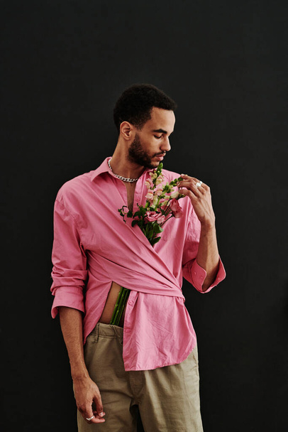 黒を背景にポーズをとりながら、ピンクのシャツの中の花の花束を嗅ぐハンサムな混合レースファッションモデル - 写真・画像