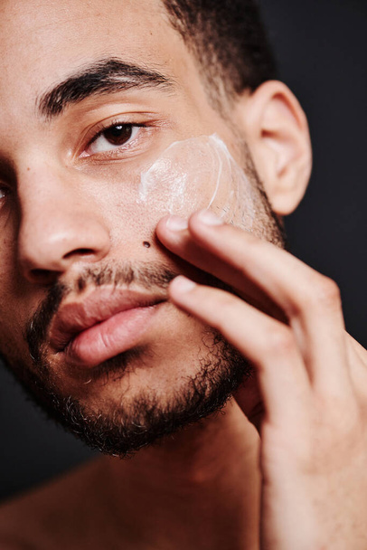 Detailní záběr vousatého mladého muže, který si nanáší krém na obličej a dívá se do kamery - Fotografie, Obrázek