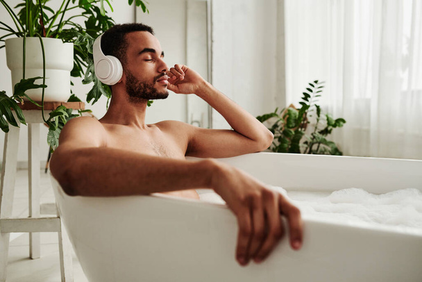 Young handsome man in wireless headphones relaxing in bath with foam and enjoying music - Photo, Image