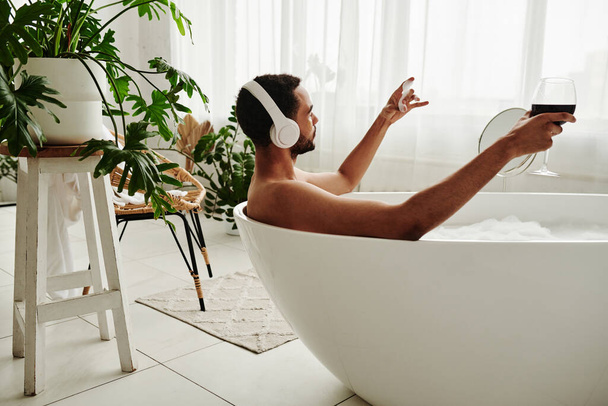 Mladý muž relaxuje ve vaně se sklenkou červeného vína a vychutnává si hudbu ve sluchátkách při lázeňské proceduře v koupelně - Fotografie, Obrázek