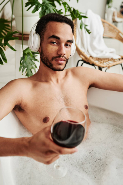 Nuoren miehen muotokuva langattomissa kuulokkeissa katselemassa kameraa viiniä juodessaan ja kylvyssä - Valokuva, kuva