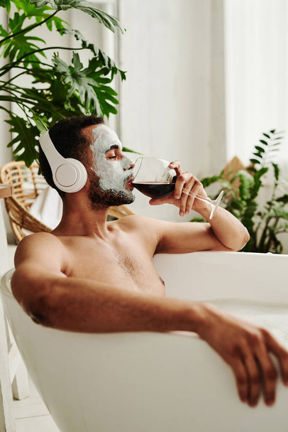 Young man in mask drinking red wine and listening to music in headphones while relaxing in bath - Foto, afbeelding