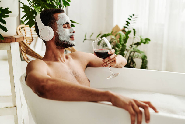 Junger Mann genießt entspannte Zeit im Badezimmer, hört Musik über drahtlose Kopfhörer und genießt ein Glas Rotwein - Foto, Bild