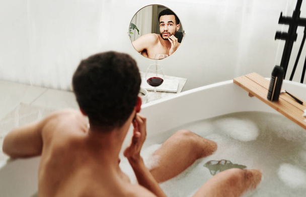 Вид спереду молодого чоловіка, який вивчає бородате обличчя у дзеркалі, сидячи у ванні
 - Фото, зображення