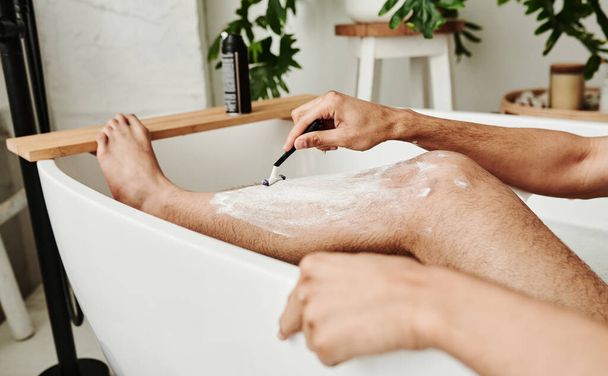 Κοντινό πλάνο του νεαρού που χρησιμοποιεί ξυράφι για να ξυρίσει τα πόδια του ενώ κάθεται στο μπάνιο. - Φωτογραφία, εικόνα