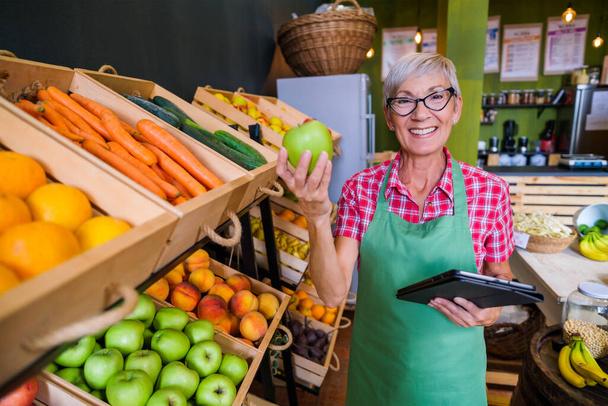 Mulher madura está trabalhando na loja de frutas e legumes Ela está examinando maçãs. - Foto, Imagem