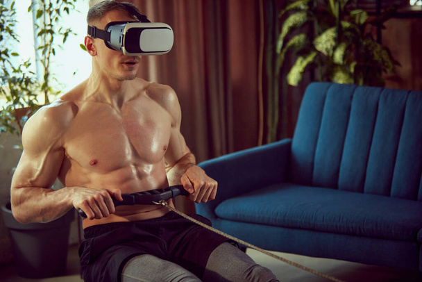 Young man with strong, muscular, relief body doing exercises with stationary rowing machine, in VR glasses at home. Online training. Sportive lifestyle, body and health care, fitness, health concept - Fotoğraf, Görsel