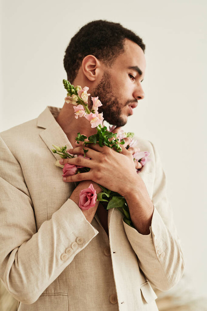 Νέοι μικτή φυλή άνθρωπος κρατώντας λουλούδια κοντά στο στήθος του και απολαμβάνοντας την ομορφιά τους απομονώνονται σε λευκό φόντο - Φωτογραφία, εικόνα