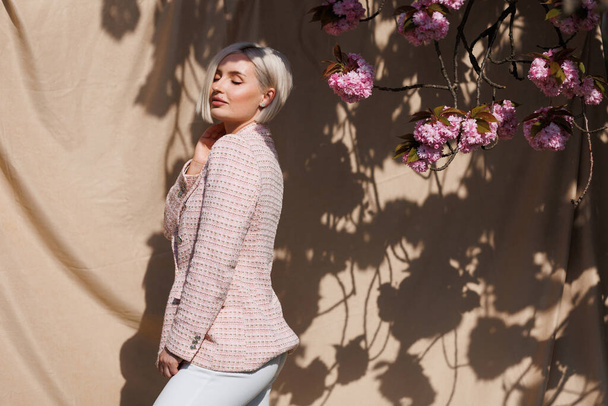 A woman with short white hair under the rays of the sun on a beige background and a branch of blooming sakura. - Photo, Image