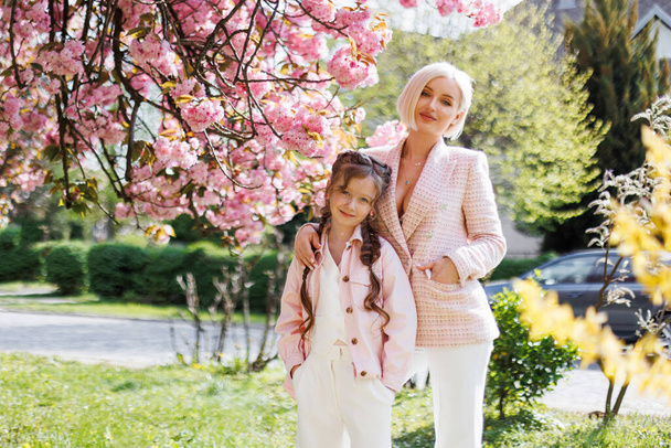 Eine Mutter und eine Tochter in pinkfarbener Kleidung stehen im Park in der Nähe eines Sakura-Baumes, der im Frühling mit rosa Blüten aufgeblüht ist. Sakura blühte auf. - Foto, Bild